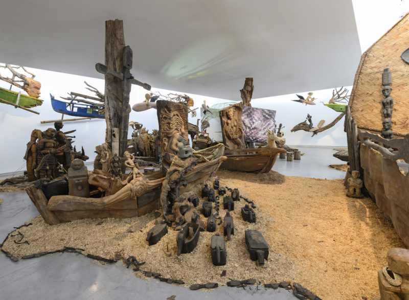 Kidlat Tahimik, Ang Ma-bagyong Sabungan ng 2 Bathala ng Hangin, A Stormy Clash Between 2 Goddesses of the Winds (WW III – the Protracted Kultur War), 2019. Wooden carved icons, ritual objects, interwoven C-print photographs, projected images, audio, mosaic, rattanbasket figurines, back-strap bamboo loom, wrought-iron launch-pads, fibreglass, root sculptures, rotten fishing boats, sawdust, bamboo fences and runo-reed fauna, dimensions variable. Installation view: Sharjah Biennial 14, 2019. Commissioned by Sharjah Art Foundation. Image courtesy of Sharjah Art Foundation 
